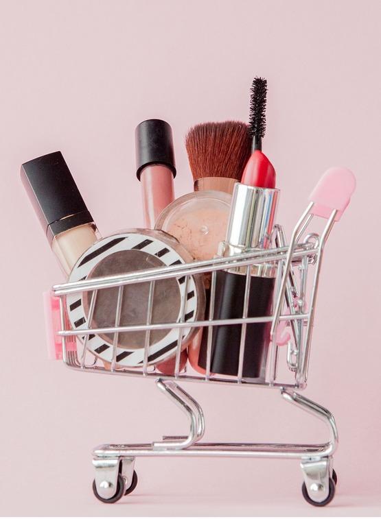 Creative concept with shopping trolley with makeup on a pink background. Perfume, sponge, brush, mascara, pencil, nail file, eye shadow, lip gloss in the basket, copy space